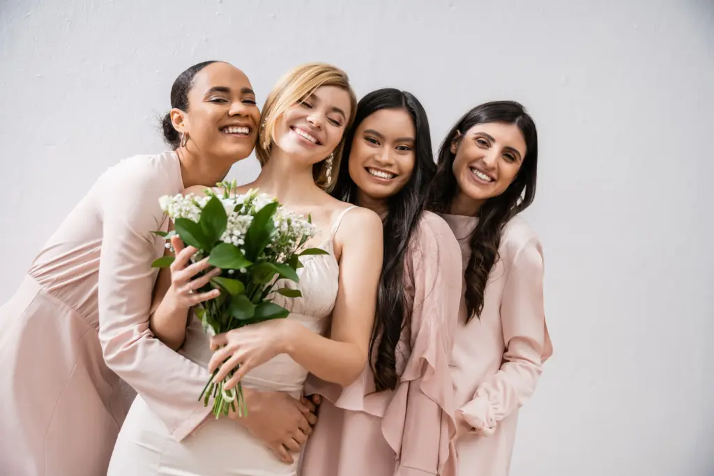Positivity, cheerful interracial bridesmaids hugging happy bride in wedding dress